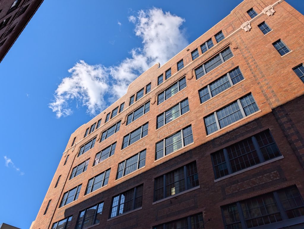 fluffy clouds emerge into blue sky from behind the top of a brown brick building