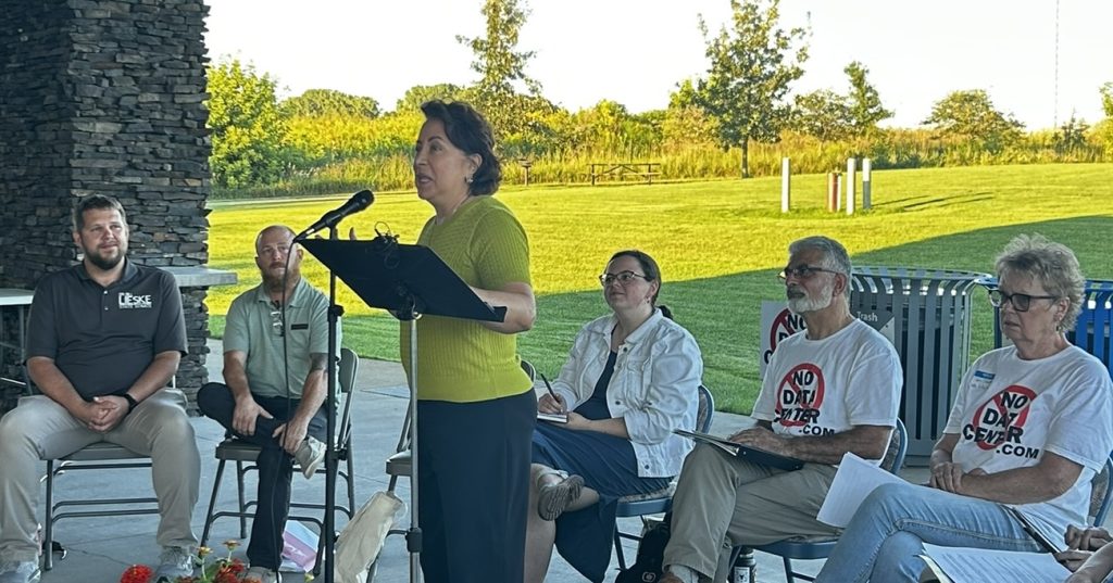 Patricia Torres Ray speaks at an outdoor community forum about hyperscale data centers, standing at a podium with a microphone under a park pavilion. Several panelists sit behind her, including Bill Lieske, Drew Roach, Sarah Mooradian, Mo Feshimi, and Cathy Johnson. Some wear white shirts that read “No Data Center.com.” A grassy park and trees are visible in the background in the late afternoon light.