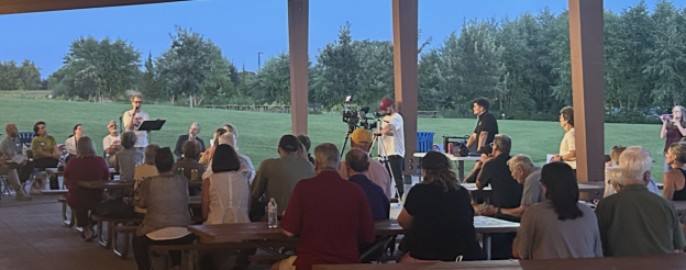 A community event taking place under a park pavilion in the early evening. A person stands at a podium speaking into a microphone while an audience of about 30 people sits on picnic benches, listening attentively. A few people operate cameras and recording equipment. The background shows a green lawn, trees, and a blue sky.