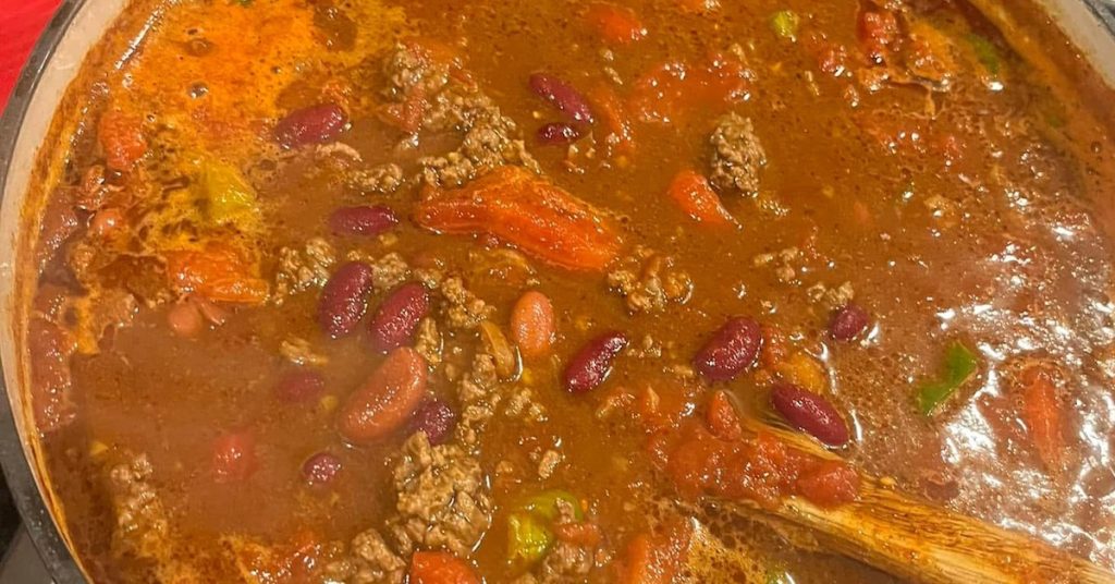A close-up, overhead view of a large, deep red cast iron Dutch oven filled with simmering chili. The chili is thick and rich, containing ground beef, red kidney beans, and chopped vegetables including chunks of red bell pepper and flecks of green pepper, with a wooden spoon resting in the pot. The pot is sitting on a black stovetop. 