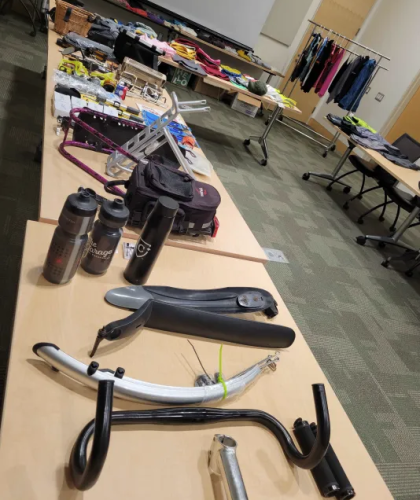 Bike fenders, water bottles, racks and other gear laid out on tables, with a rack of clothes in the background.