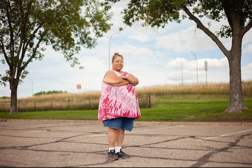 A woman standing in a parking lot, behind her are two trees and a field. 