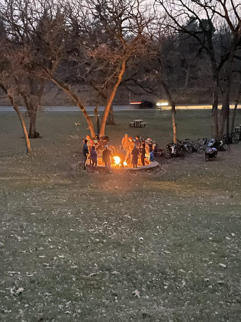 People surrounding a bonfire near dusk in a park. Parked bikes in the trees to the right.