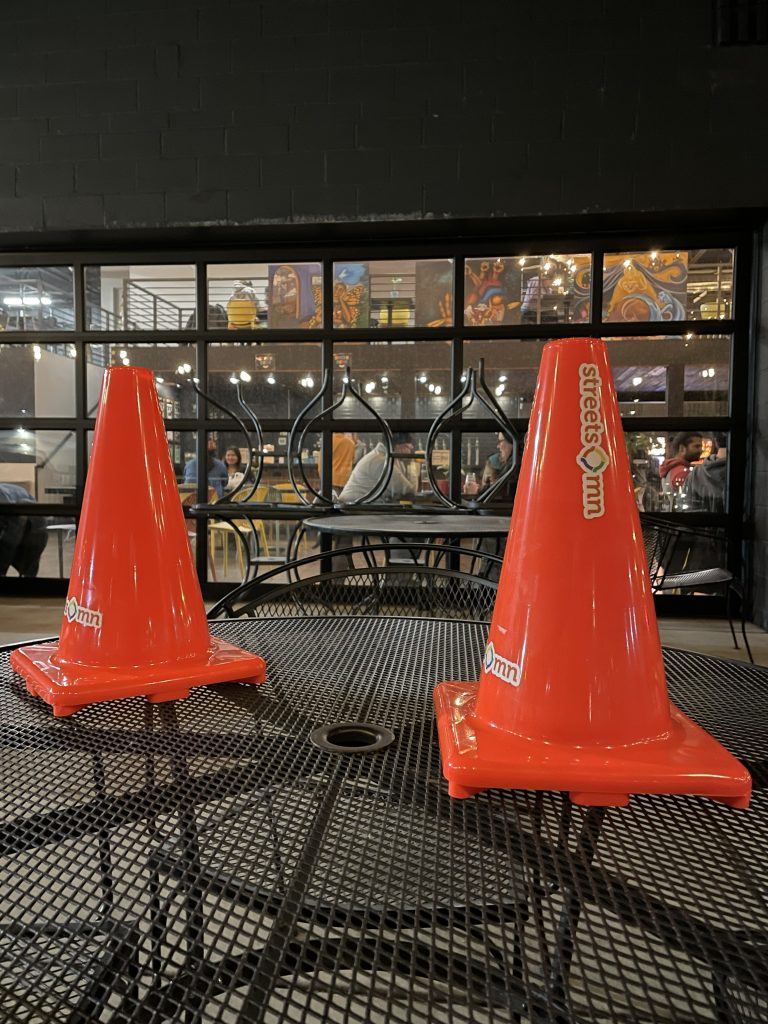 Two orange traffic cones with Streets.mn stickers on them sitting on a table on a patio