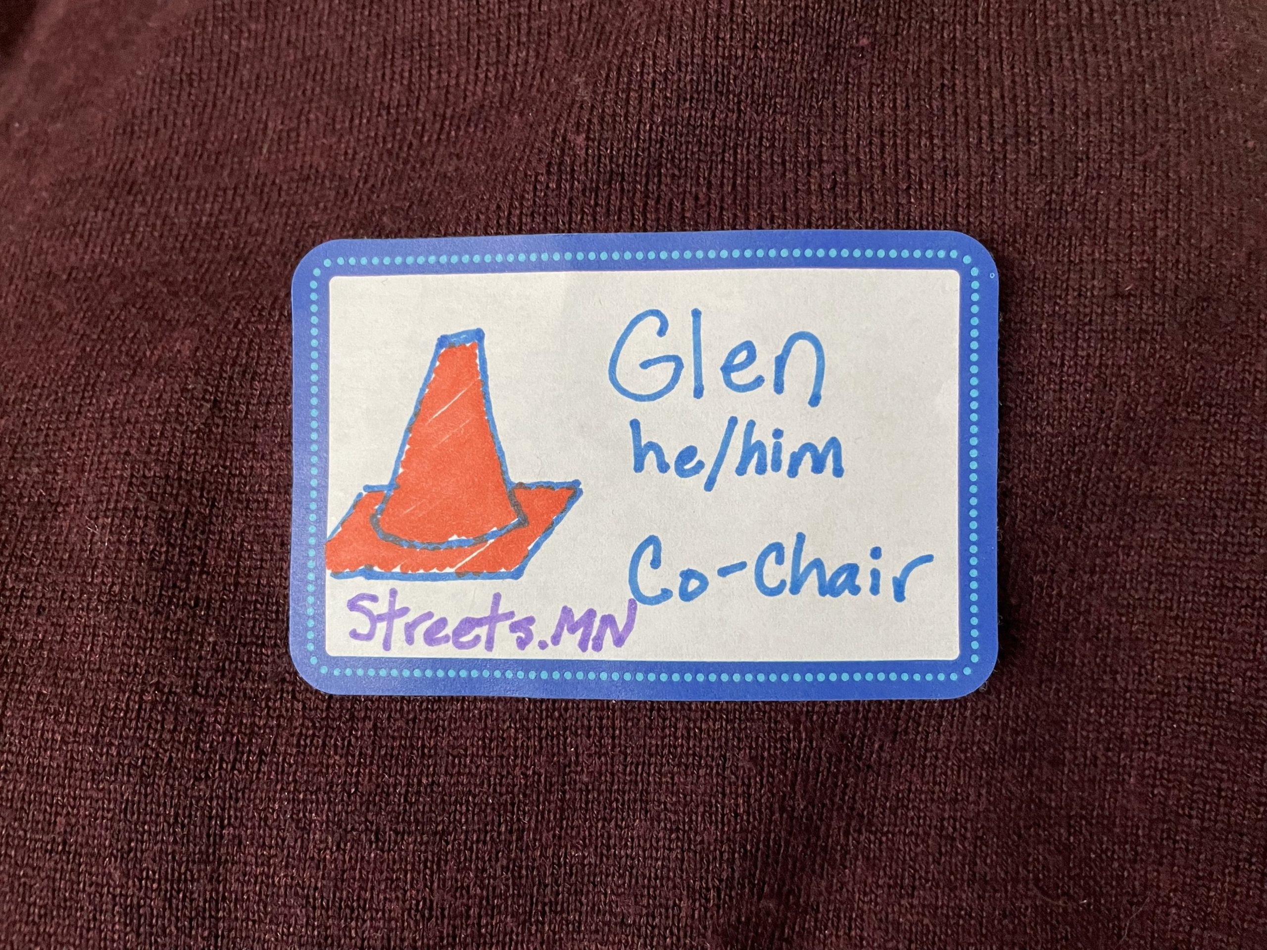 Nametag with a drawn orange traffic cone and words Glen, he/him, Co-Chair, and Streets.mn