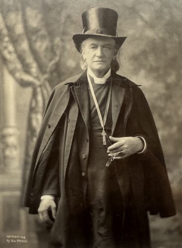 Black and white photograph of Bishop Whipple in 1898. He is wearing a pectoral cross around his neck with an overcoat draped over this shoulders and a top hat on his head.