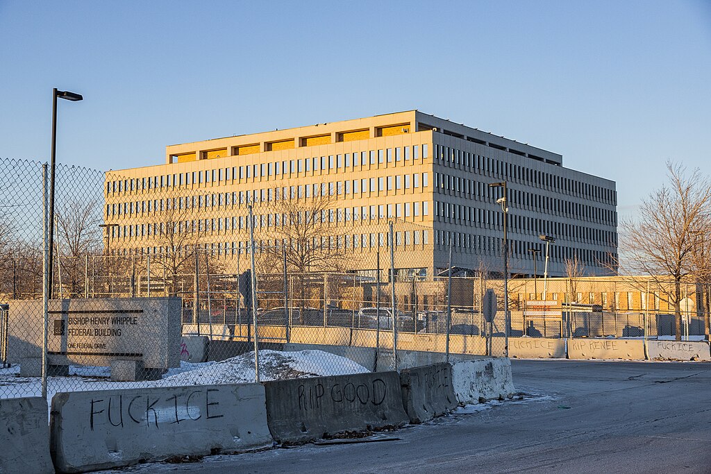 Photograph of the Whipple Federal building with chain-link fence and concrete barricades and snow around it. Anti-ICE messages are written on the barriers.