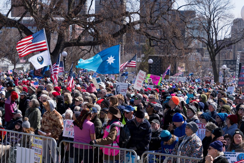 Large crowd of demonstrators behind a fence at No Kings 3 with U.S. flags, a Minnesota state flag, and a Minnesota resistance flag. Photo: Conrad Lange Zbikowski