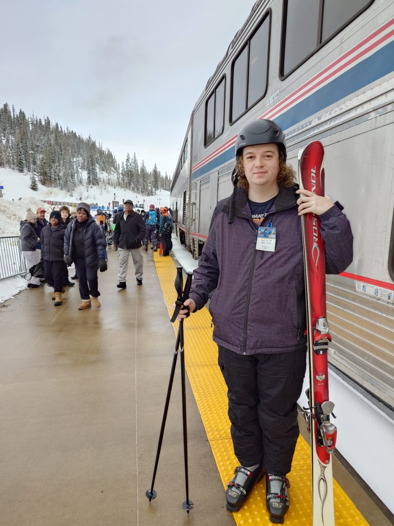 Ian Gaida standing with downhill skis next to an Amtrak train at Winter Park station