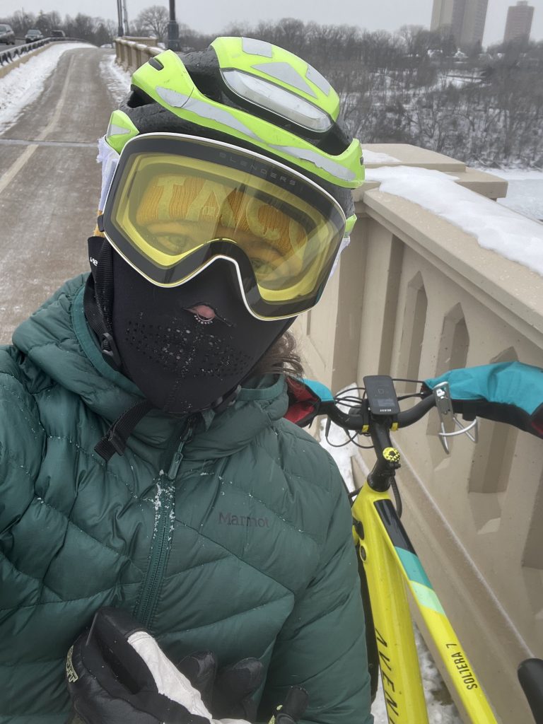 angela olson wearing goggles, helmet, and face cover standing next to a bike in the winter