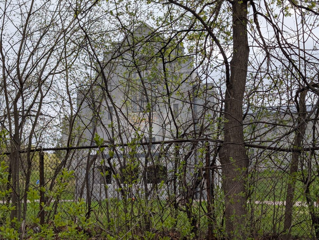 cathedral-like limestone building screened by overgrowth and a chain-link fence