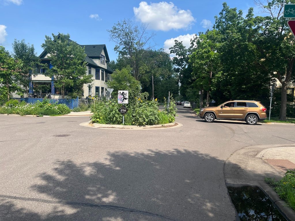 A car drives by a planted traffic circle at a residential intersection.