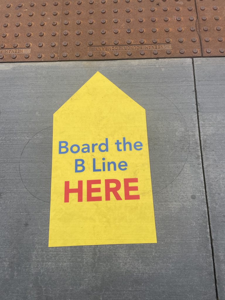 Painted sidewalk sign: Board the B Line here.