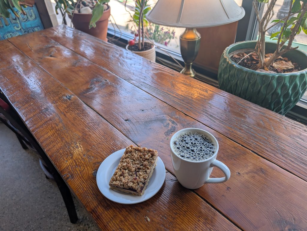 wooden table with coffee and a bar, behind which are potted plants and a lamp followed by windows
