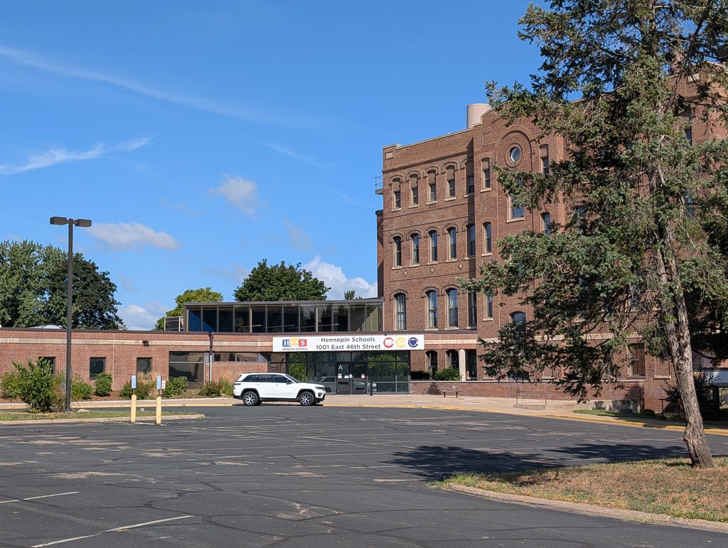 the same four-story brick building as shown earlier joined to a one-story section with a Hennepin Schools sign