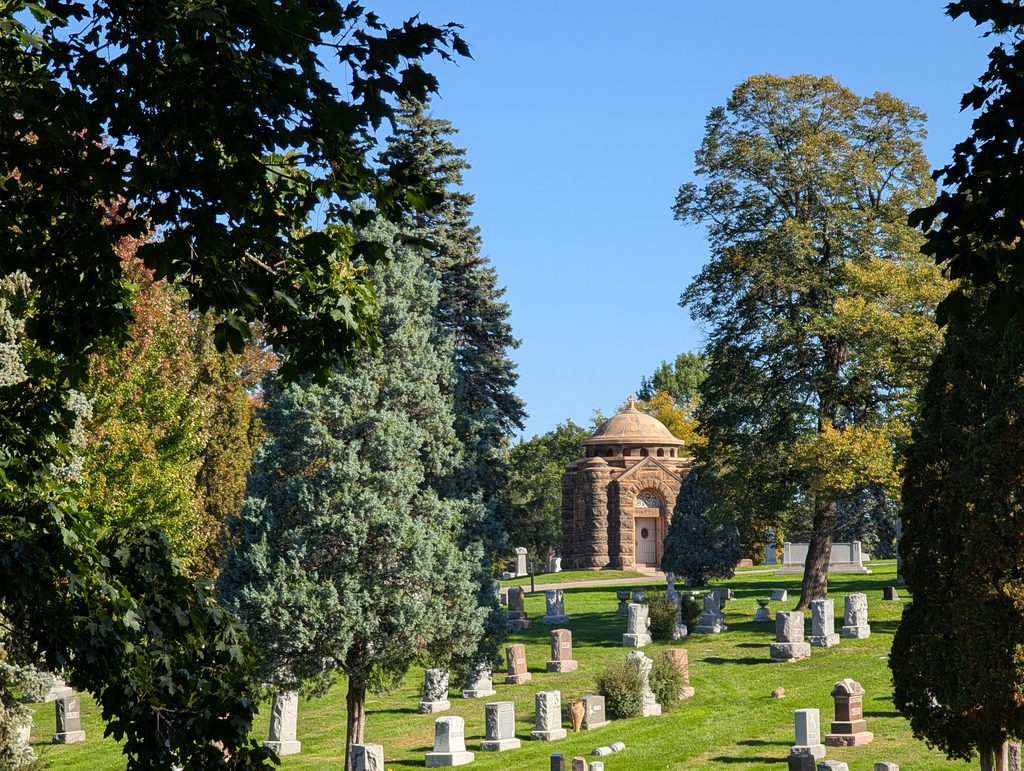 grassy cemetery with gravestones in foreground amidst trees and in the background a Richardsonian Romanesque mausoleum