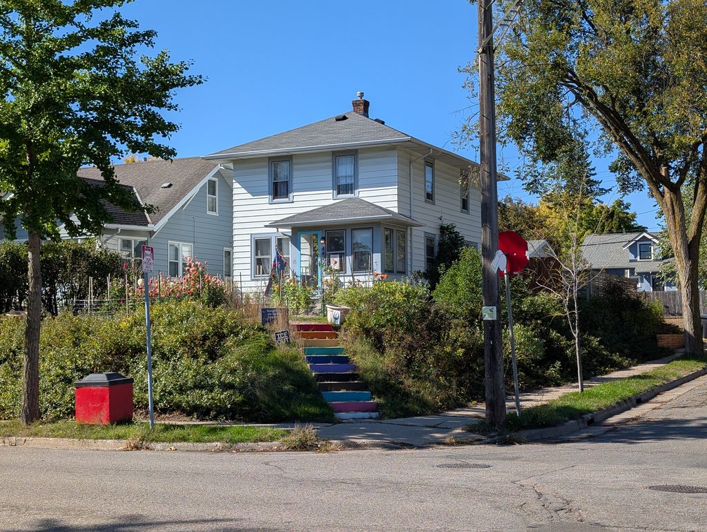 white two-story house on corner lot elevated above street level and reached via multicolored stairs from the sidewalk corner