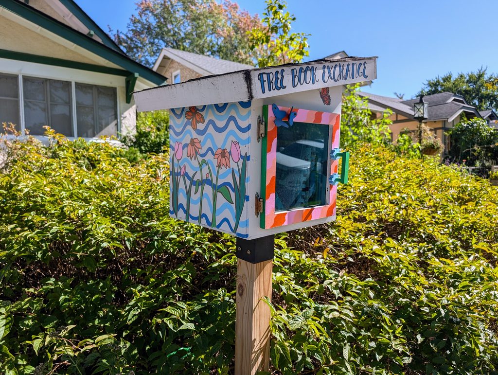 free book exchange box on post is painted on the side with flowers against a background of blue waves and around the front door in diagonal stripes of pink and orange with a green handle and blue butterlies