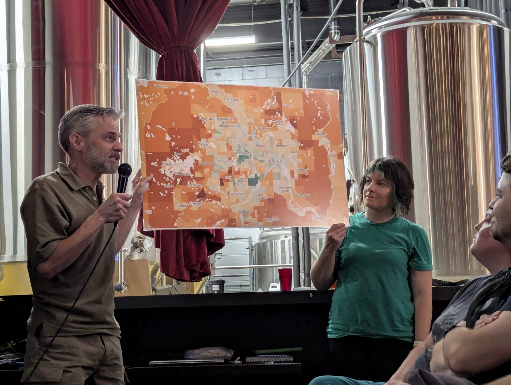Two people holding up a large map for the crowd to see. The map shows the Twin Cities metro area, color coded with the city centers generally shaded more green, and the outer suburbs generally shaded more orange and red.